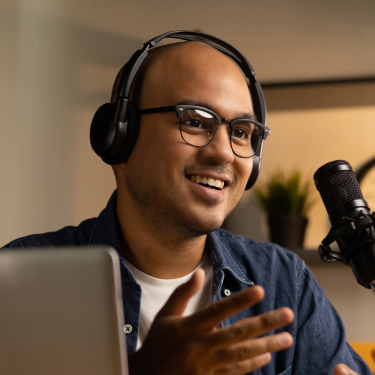Smiling young man wearing headphones with a laptop and microphone in front of him.