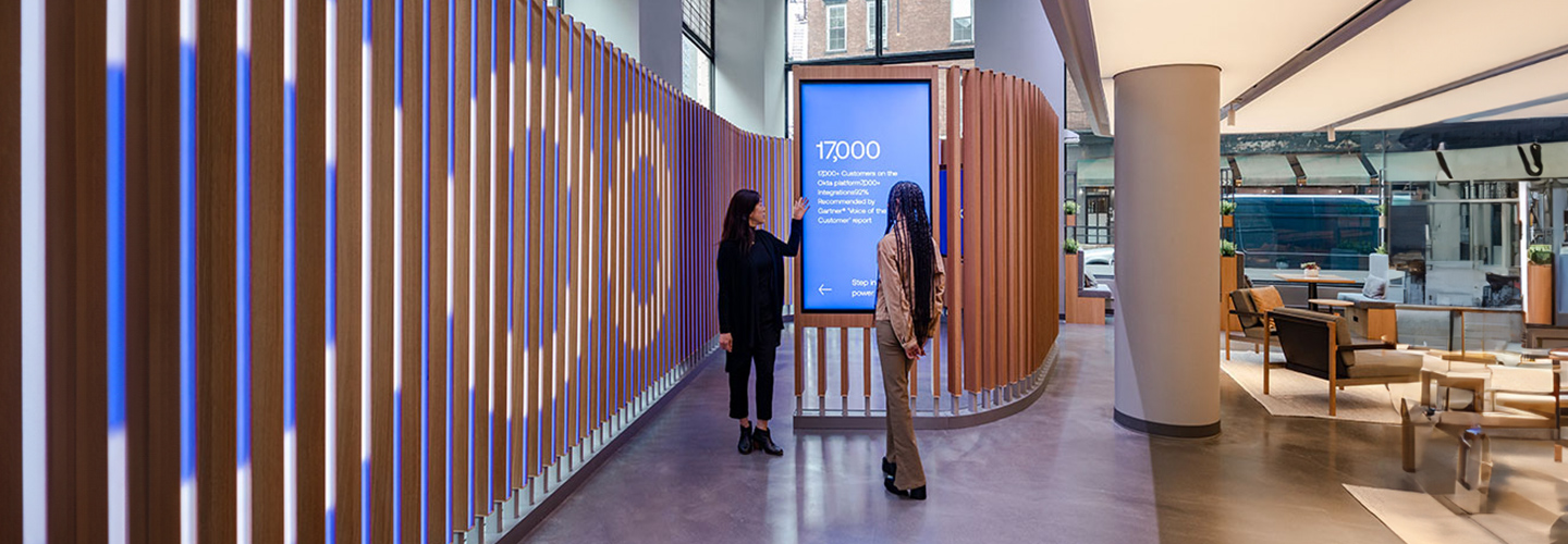 Two people standing in front of a digital screen in the New York Okta office.